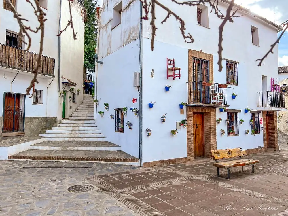 a cute square with wood carved benches and potplants and colorful chairs hanging on the whitewashed walls in Parauta near Ronda - white villages Spain