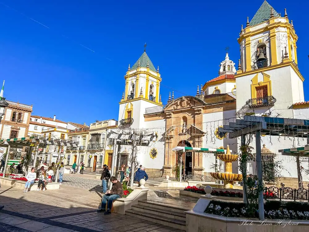 a quaint square with a fountain in front of a white and yellow church with two towers - what to see in Ronda Spain