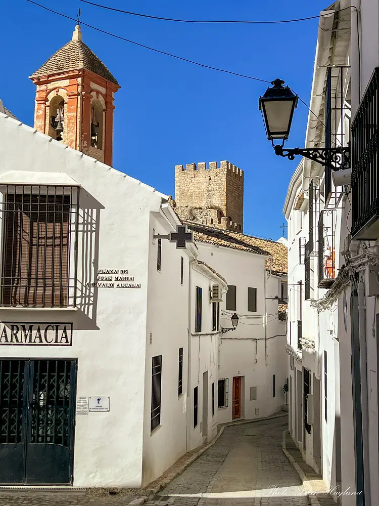 Streets of Zuheros Cordoba with whitewqashed houses and a red church tower and a watch tower seen above the houses.